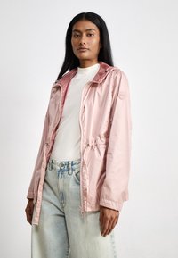 Chaqueta ligera de color rosa claro, con cintura ajustable y capucha, que presenta una textura suave y cierre frontal con cremallera. Combinada con jeans de mezclilla claros.