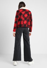 Rød og sort ternet cropped jakke lavet af uldlignende materiale med hvid falsk pelskrave. Sorte jeans med vide ben og hvide sneakers.