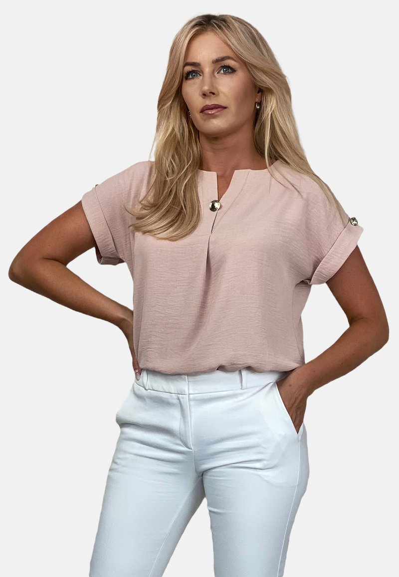 Różowa bluzka krótki rękaw z okrągłym dekoltem i jednym złotym guzikiem przy kołnierzy, w połączeniu z białymi dopasowanymi spodniami.