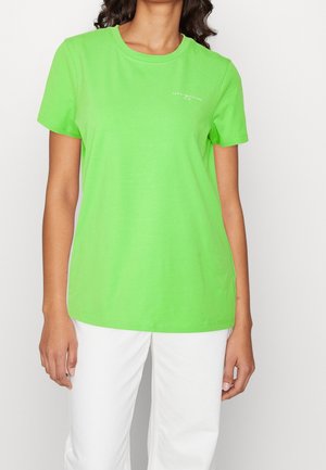 Kvinde iført en knaldgrøn kortærmet t-shirt med lille hvid tekstlogo på venstre bryst, parret med hvide bukser.