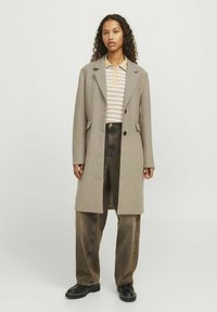Abrigo de lana beige con solapas en pico y bolsillos laterales, llevado sobre una camisa de rayas con cuello, combinado con jeans marrones holgados y botines negros.