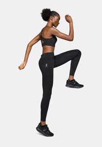 Czarny stanik sportowy z wycięciami, w połączeniu z czarnymi legginsami z logo. Model w ruchu, również w czarnych butach sportowych.