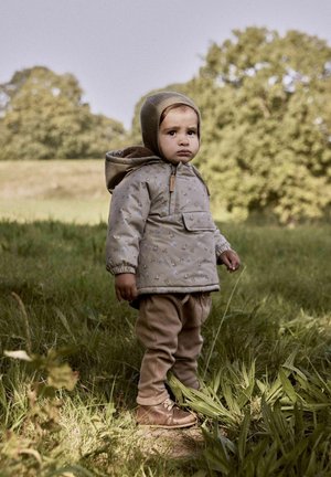 Cappotto imbottito marrone chiaro con motivi floreali, pantaloni beige abbinati e scarpe marroni. Modella indossa un cappello di maglia, in piedi sull'erba.