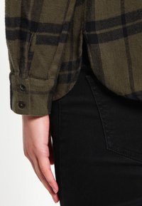 Olivgrön rutig skjorta med mörkblå linjer och knäppte manschetter, bärs över svarta jeans och visar en strukturerad tyg och skräddarsydd passform.