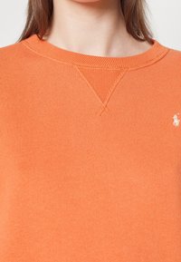 Osoba v oranžovém crewneck svetru s texturovaným švem a malým bílým vyšitým logem na pravé straně hrudi.