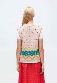 Chaleco sin mangas de punto en rosa claro con puntos rojos. Presenta patrones geométricos en verde y naranja en el dobladillo. Cuello y sisas acanalados.