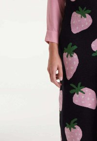 Suéter de punto en negro con grandes patrones de fresas rosas y hojas verdes. Textura suave, mangas largas y una camiseta rosa debajo.