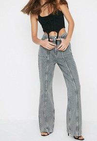 Mujer vistiendo un top corto negro y jeans gris de pierna ancha de alta cintura con detalles de costura, sosteniendo la cintura, de pie contra un fondo blanco.