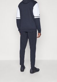 Marineblauer Kapuzenpullover mit weißen Akzenten und Streifen, kombiniert mit passenden marineblauen Jogginghosen. Schwarze Sportschuhe mit strukturierten Sohlen runden das Outfit ab.