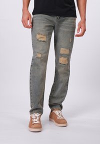 Jean bleu clair déchiré avec des bords effilochés, présentant plusieurs trous repris. Associé avec des baskets en daim beige et des lacets blancs.