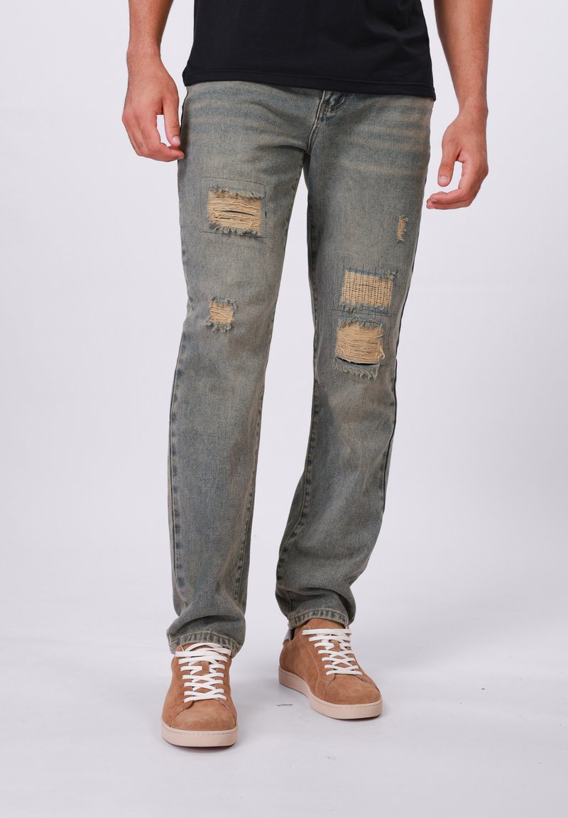 Jean bleu clair déchiré avec des bords effilochés, présentant plusieurs trous repris. Associé avec des baskets en daim beige et des lacets blancs.