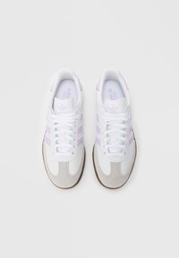 Une paire de baskets Adidas blanches avec des rayures violettes claires et des embouts en daim beige, présentée de dessus sur un fond blanc.