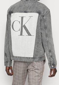 En person står klädd i en grå denimjacka med en stor vit panel med "CK" tryckt på ryggen, tillsammans med rutiga byxor.