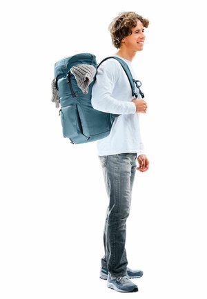 Blauer Rucksack mit mehreren Fächern, Gurten und einem gestrickten Gegenstand, der oben drapiert ist; getragen von einer Person in lässiger Kleidung.