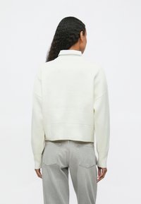 Γυναίκα με μακριά μαύρα μαλλιά φορώντας ένα λευκό πουλόβερ πάνω από ένα πουκάμισο και ανοιχτό γκρι τζιν, στραμμένη πίσω με φόντο λευκό.