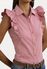 Ljusrosa ärmlös blus med volangdetaljer på axlarna, knappstängning och en figursydd design, kombinerad med blå denim och ett brunt bälte.