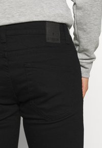 Jeans de mezclilla negra con un diseño ajustado, que incluyen un bolsillo trasero y un parche de cuero en la cintura. Llevados con una camiseta de manga larga gris.