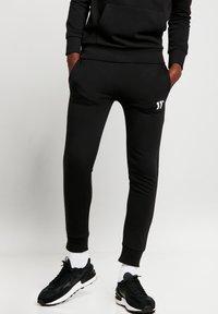 Pantalones de chándal negros hechos de una tela suave con un corte ajustado, que cuentan con bolsillos laterales y un pequeño logo blanco cerca de la cintura.