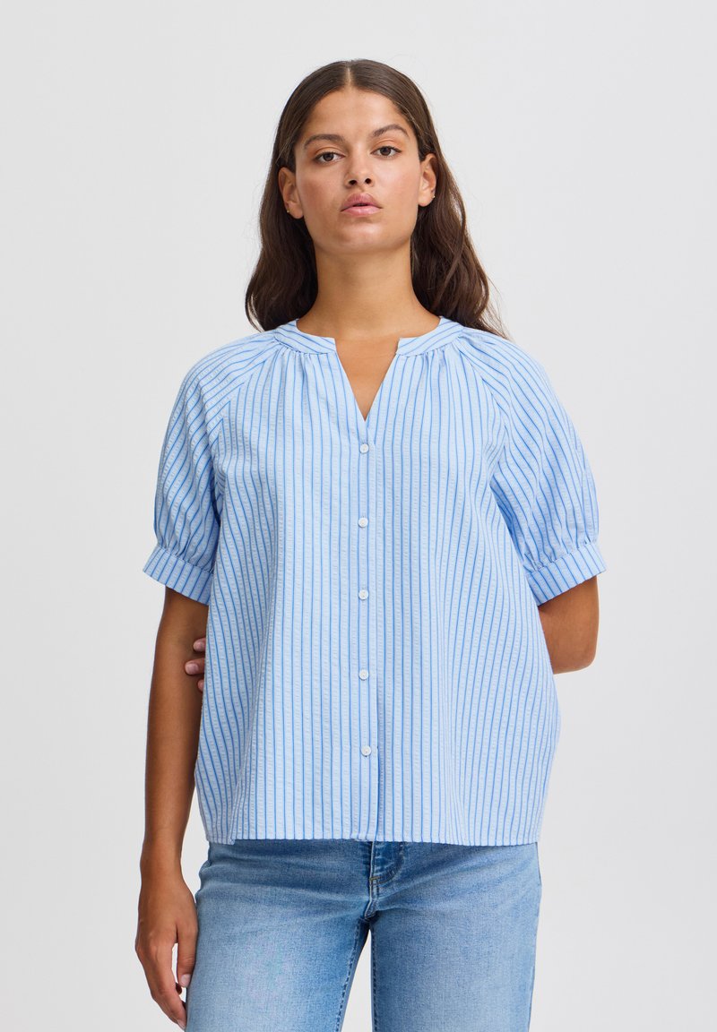 Blusa a righe azzurro chiaro con maniche a sbuffo corte, chiusura frontale a bottone e scollatura rotonda. Il tessuto morbido e le righe verticali creano una vestibilità rilassata.