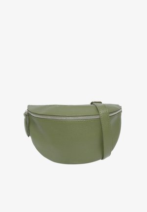 Jeune femme portant un haut noir et un pantalon noir avec un grand sac bandoulière vert porté en travers de la poitrine, souriante et regardant de côté.