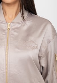 Donna che indossa una giacca bomber di raso beige chiaro con cerniera dorata e la scritta "Bomber Original" ricamata sul petto.