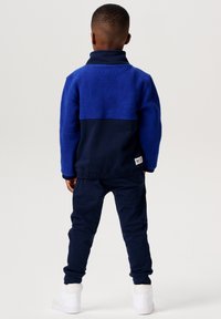 Marineblauwe fleece pullover met hoge kraag, gecombineerd met bijpassende marineblauwe broek en witte sneakers. Zachte textuur, casual ontwerp.