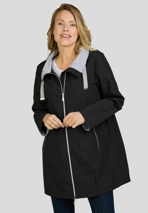 Femme portant une veste noire zippée avec capuche grise et cordons, souriant les mains près de la fermeture éclair, sur un fond clair uni.