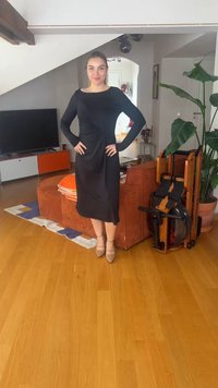 Femme en robe noire et chaussures beiges se tient les mains sur les hanches dans un salon avec un sol en bois, un canapé orange, une plante et un rameur.