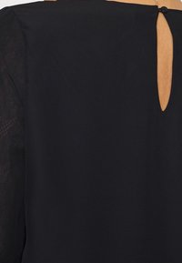 Blouse noire en tissu uni avec détails en dentelle sur les manches et une ouverture en forme de goutte dans le dos, fermée par un bouton.