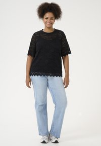 Black lace top with short, scalloped sleeves and hem, paired with light blue jeans. Model stands on a plain background, smiling.