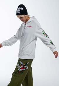 Sudadera blanca con cremallera y bordado negro y rojo, de corte holgado. Pantalones verde oliva con bordado colorido de un pájaro y una pancarta en el costado.