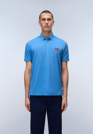 Mand stående iført lyseblå kortærmet polo med norsk flagemblem og mørke bukser mod en ensfarvet, lys baggrund.