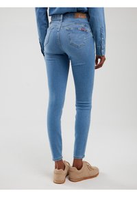 Isik, kes kannab helesiniseid kitsaid teksaseid ja beeže tossusid, seisab seljaga kaamerasse tavalise valge tausta ees.