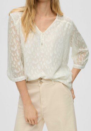 Femme portant un chemisier blanc transparent à motifs avec des manches trois-quarts et un pantalon beige taille haute à double boutonnage.