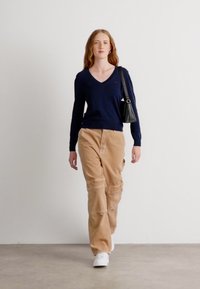Maglione blu a scollo a V, pantaloni cargo beige con dettagli di cucitura e sneakers bianche. La modella porta una piccola borsa nera.