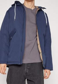 Giacca piumino blu navy con cappuccio, dotata di foderatura beige, chiusura frontale con zip e bottoni a pressione. Indossata sopra una maglietta grigia e jeans scuri.