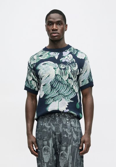 Πλεκτό πουλόβερ με κοντά μανίκια σε ναυτικό μπλε χρώμα με ζωηρό λουλουδένιο σχέδιο σε αποχρώσεις του πράσινου και του μοβ. Συνδυάζεται με υφασμάτινο γκρι παντελόνι με σχέδιο και υφή.