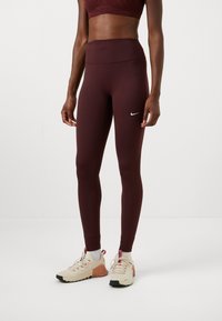 Burgunderfargede høytlivs leggings med en glatt tekstur og en liten hvit Nike-logo på låret, kombinert med beige og oransje treningssko.