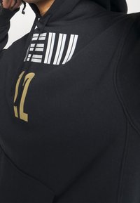 Felpa nera con finitura testurizzata, presenta stampe grafiche bianche e dorate, comprese strisce e il numero 12. Visibili le stringhe della cappuccio regolate.