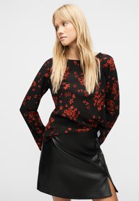 Femme aux longs cheveux blonds portant un haut noir à manches longues avec un motif floral rouge et une jupe en cuir noire fendue sur le côté, regardant vers sa droite.