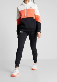 La sudadera con capucha presenta un diseño de bloques de color en naranja, blanco y negro; se combina con joggers negros. Ambos están hechos de una tela suave.