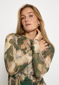 Garais piedurkņu tops ar caurspīdīgu kamuflāžas rakstu zaļā un bēša krāsā. Iezīmē apļa kakla izgriezumu un pieguļošu siluetu.