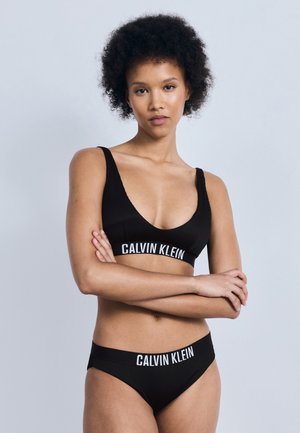 Vrouw met krullend haar, gekleed in zwarte Calvin Klein onderkleding, staand met gekruiste armen tegen een eenvoudige grijze achtergrond.