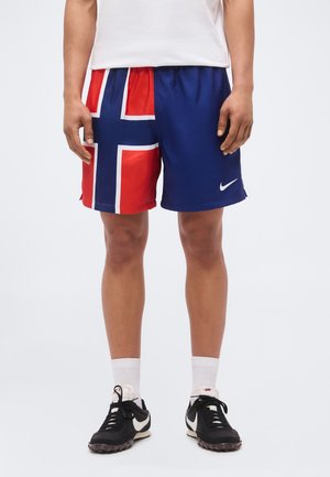 Mann med blå og røde shorts med hvitt korsdesign, hvite sokker, svart-hvite Nike-sneakers, stående på ensfarget bakgrunn.