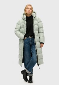 Polstret, mellemlangt frakke i lys mintgrøn, med hætte, trykknaplukninger og sidelommer. Båret over en mørk sweater og blå jeans.