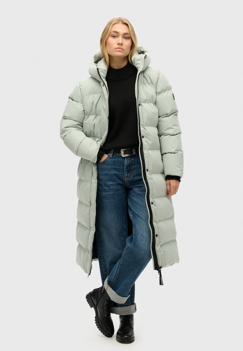 Polstret, mellemlangt frakke i lys mintgrøn, med hætte, trykknaplukninger og sidelommer. Båret over en mørk sweater og blå jeans.