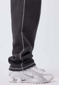 Persona vistiendo pantalones de chándal oscuros y zapatillas Nike blancas con suelas distintivas tipo resorte, de pie contra un fondo claro y liso.