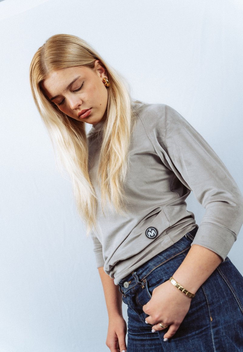 Grijs top met lange mouwen en een ronde halslijn met een logo ter hoogte van de heup, gecombineerd met donkerblauwe denim jeans en een gouden armband.