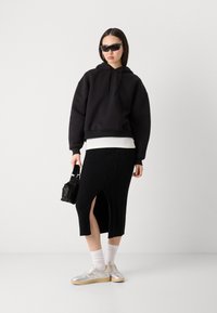 Sweat-shirt noir oversize avec une poche frontale, superposé sur un haut blanc. Jupe noire à côtes avec fente sur le côté et motif en tricot à câble. Baskets argentées.