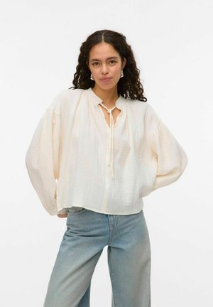 Blouse à manches longues crème clair en tissu texturé avec un col rond et une fermeture à nouer, associée à un jean bleu taille haute.